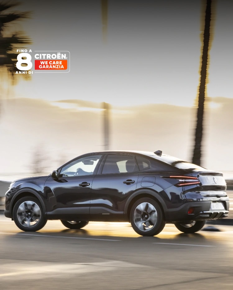 Citroën ë-C4 X in marcia lungo una strada costiera al tramonto con il badge della copertura Citroën 8 anni esposto